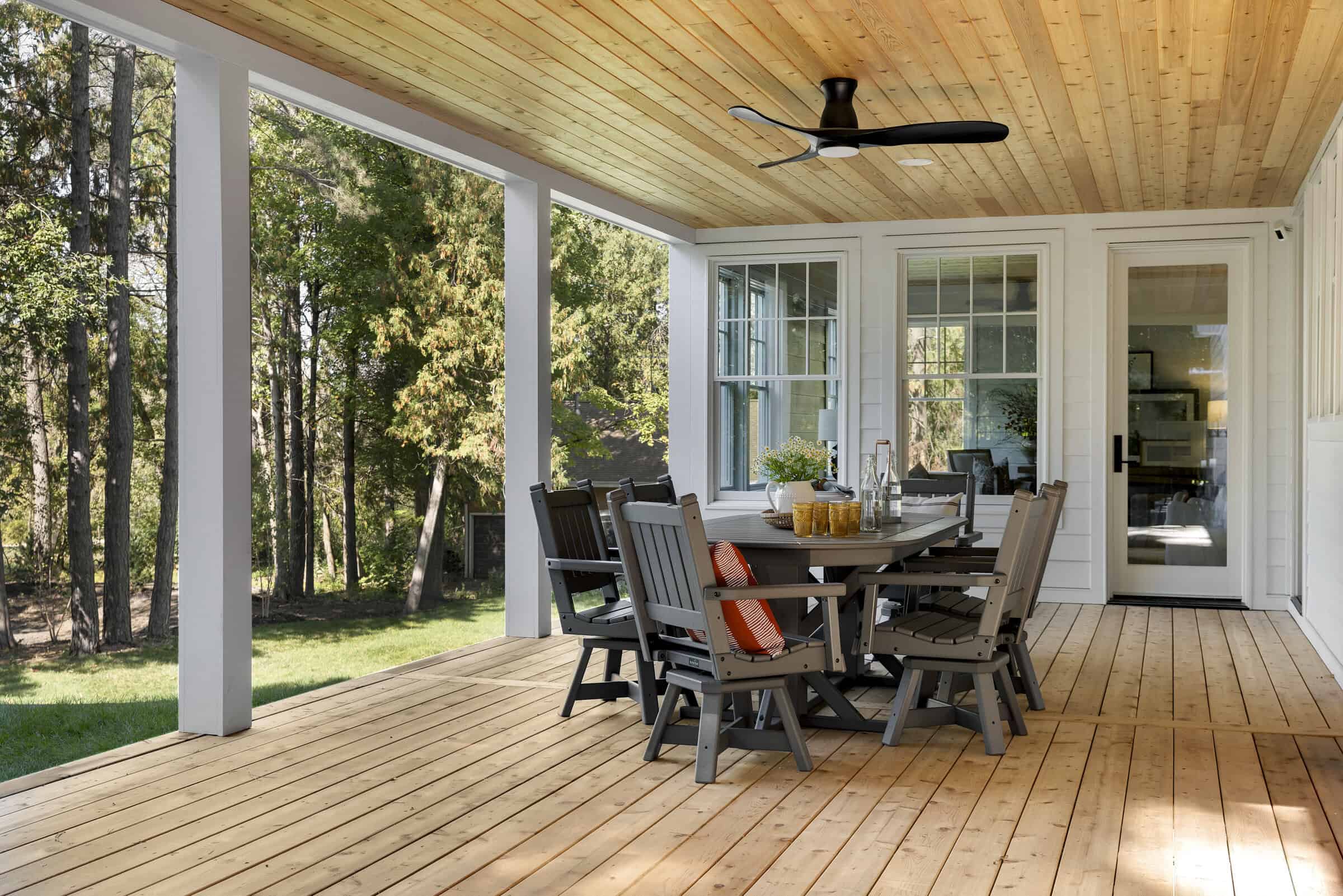 Covered wooden porch with dining table, ceiling fan, and trees